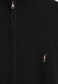 Svart stickad zip-up-tröja med ribbad krage, slät textur och en liten broderad logotyp i en ljusare nyans på vänster sida.