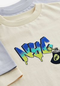Artimu du prisiūto megztinio, vienas kreminių spalvų su mėlynais ir žaliais graffiti stiliaus "NYHC" ir purškimo dažų skardinės dizainu, o po juo šviesiai pilkas megztinis.