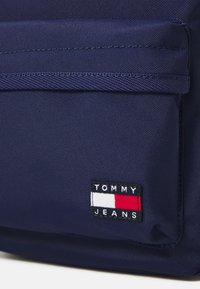 Tengerészségszínű vászontáska elülső zsebbel és egy fekete folttal, rajta a "TOMMY JEANS" felirattal és piros, fehér, valamint kék vízszintes csíkokkal. Texturált felület.