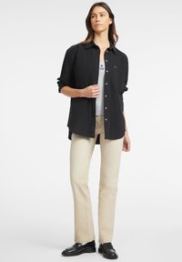 Camicetta nera allacciata, realizzata in cotone, con taschino sul petto, indossata sopra una canotta grigia, abbinata a jeans beige a gamba dritta e mocassini neri.