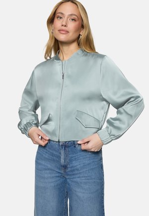 Femme portant une veste bomber en satin bleu clair avec fermeture éclair et poches frontales, associée à un jean bleu taille haute, les mains ajustant l'ourlet de la veste.