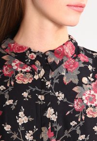 Donna che indossa una camicia nera con colletto, decorata con stampa floreale rossa, rosa e beige, che mostra il profilo del collo e della parte inferiore del viso senza gli occhi.