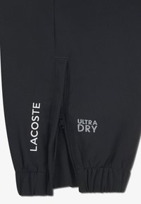 Black athletic pants with a zippered pocket, "LACOSTE" logo in white, and "ULTRA DRY" text in gray on the left side. Textured fabric.