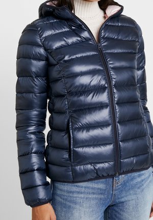 Femme portant une doudoune brillante bleu marine zippée avec une capuche, par-dessus un pull blanc à col roulé, associée à un jean bleu clair.