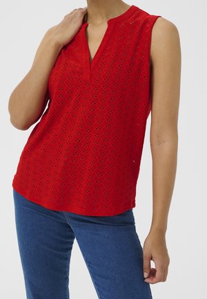 Blusa roja sin mangas con diseño de ojales y escote en V combinada con vaqueros azules, modelo posando con una mano sobre el hombro y otra hacia abajo.