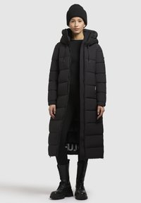 Melna pūkaina jaka ar garu, iešūtu dizainu, kapuci un priekšējo rāvējslēdzēju. Valkāta ar piegulošu melnu apģērbu un izturīgām melnām šņorējamām zābakiem.