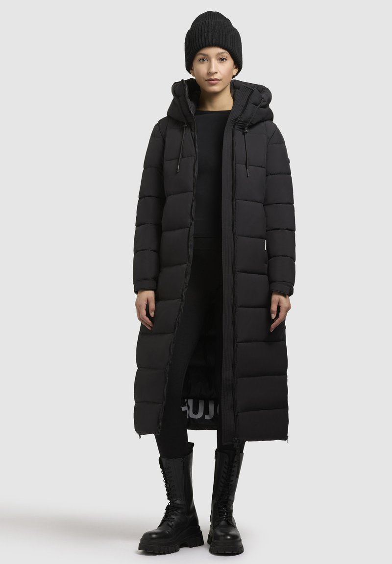 Melna pūkaina jaka ar garu, iešūtu dizainu, kapuci un priekšējo rāvējslēdzēju. Valkāta ar piegulošu melnu apģērbu un izturīgām melnām šņorējamām zābakiem.