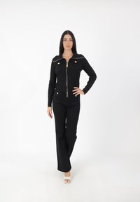 Schwarze gerippte Jacke mit goldenen Reißverschlüssen und Knöpfen, kombiniert mit schwarzen Schlaghosen. Das Modell trägt transparente Absatzsandalen vor einem weißen Hintergrund.