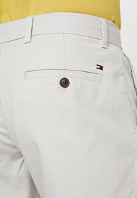 Pantaloni corti grigi chiari in cotone misto; presentano una tasca posteriore con bottone e un piccolo logo decorativo in rosso, bianco e blu.