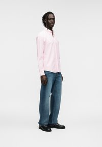 Chemise rose clair à boutons, manches longues, associée à un jean en denim bleu à jambes droites et des chaussures en cuir noir. Design simple et classique.