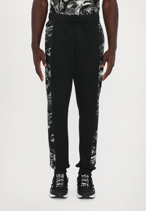 Black sweatpants with a patterned side panel, elastic waistband, and drawstring. Paired with black and white sneakers featuring text.