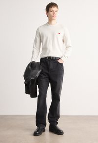 Pull long à manches longues crème claire, pantalon en denim noir, veste en cuir noir drapée sur le bras et bottes noires. Design simple, couleurs unies.