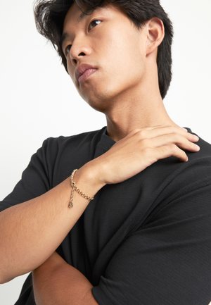 Persona joven con camiseta negra tocándose el hombro con una mano, mientras el otro brazo cruzado lleva una pulsera de cadena dorada.