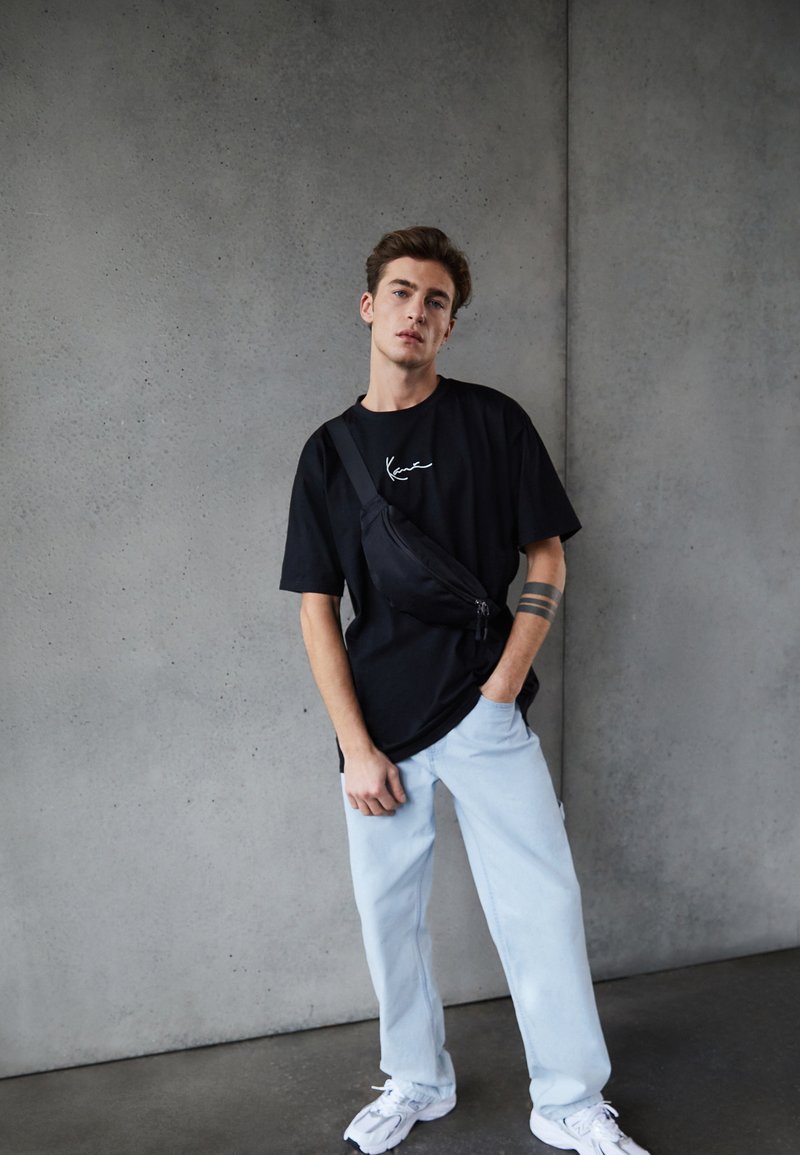 Maglietta nera a maniche corte con logo bianco, jeans azzurri larghi, borsa a tracolla nera e sneaker bianche. Texture lisce; design casual.