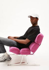 Chair with plush, pink upholstery, modern design, metal base. Person seated, wearing a black shirt and white cap, gray jeans, and sneakers.