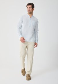 Light blue long-sleeve linen shirt with a henley collar and single chest pocket, paired with cream-colored trousers and brown slip-on shoes.