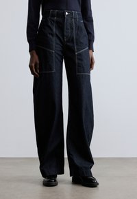 Tamnoplavene široke traperice s kontrastnim svijetlim šavovima, visokim strukom i velikim prednjim džepovima. Glatka tekstura, minimalni metalni dijelovi.