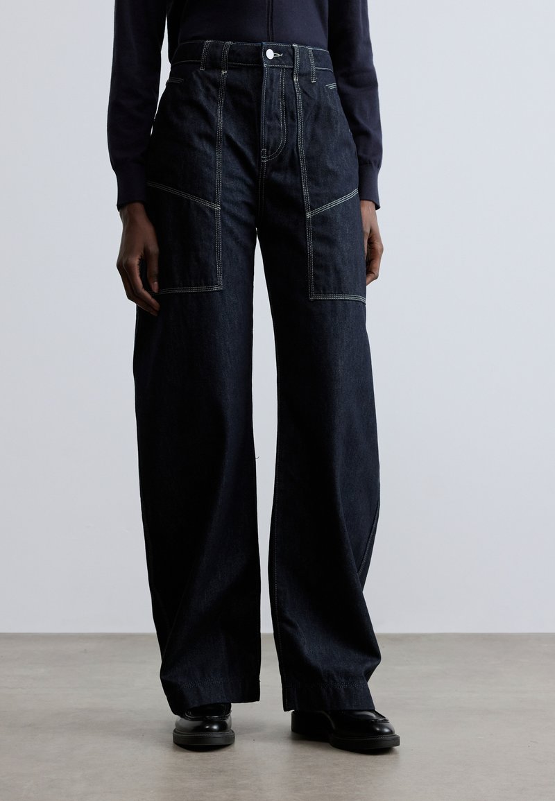 Tamnoplavene široke traperice s kontrastnim svijetlim šavovima, visokim strukom i velikim prednjim džepovima. Glatka tekstura, minimalni metalni dijelovi.