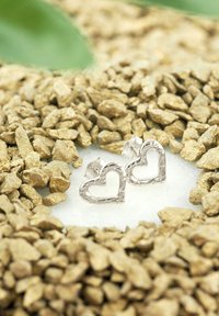 Silver hjärtformade örhängen med en utskärd design, som har en texturerad yta. Visas bland små gyllene stenar.