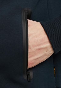 Marineblauwe stoffen ritszak met een zwarte rits en getextureerde randen, waarbij een hand de ritstrekkker vasthoudt op een donkere achtergrond.