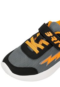 Szare i czarne trampki z pomarańczowymi sznurowadłami, pomarańczowym wzorem błyskawicy oraz czarnym zapięciem ustnym z pomarańczowym napisem na białej podeszwie.