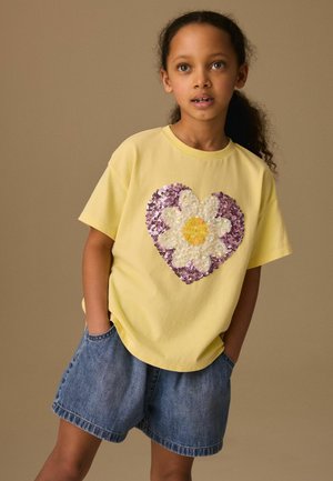 Pige med krøllet hår iført en gul T-shirt med et hjerte i pailletter og margueritdesign, hænderne i lommerne, og blå denimshorts.