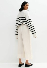 Gestreepte zwart-witte gebreide trui, losvallende crèmekleurige broek en zwarte ballerina's, gezien vanuit de achterkant tegen een neutrale achtergrond.