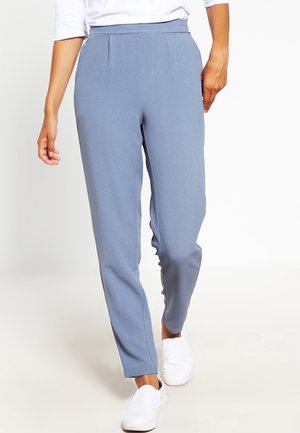Pantalones ajustados de color azul claro con una textura suave, que presentan pliegues delanteros y una cintura ajustada, combinados con zapatos blancos tipo slip-on.