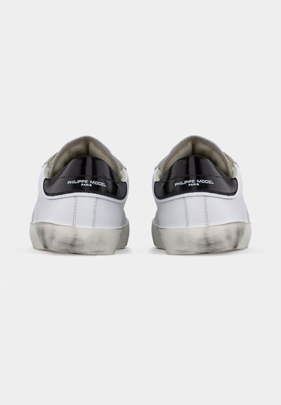 Paire de baskets basses blanches avec des languettes noires au talon et le texte « Philippe Model Paris », vues de dos avec des semelles légèrement usées.