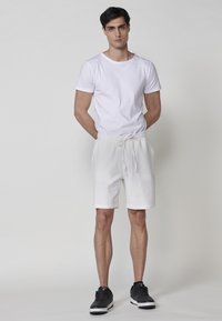 Giovane uomo in piedi con le mani dietro la schiena, indossando una t-shirt bianca, pantaloni corti bianchi con texture e coulisse, e scarpe da ginnastica nere su uno sfondo neutro.