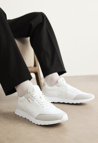 Hvite joggesko med lysegrå detaljer, glatte og teksturerte materialer, med snøring og en liten rød logo på siden.