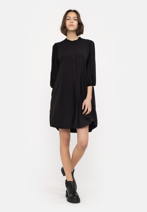 Robe noire avec une patte de boutonnage, un col rond et des manches bouffantes. Fabriquée en tissu lisse, elle présente une coupe ample, à mi-genoux, avec des poches latérales.