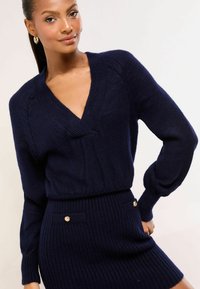 Námornícky modrý pletený sveter s hlbokým výstrihom do V a rebrovanými manžetami, v kombinácii so zodpovedajúcou rebrovanou sukňou s ozdobnými zlatými gombíkmi.