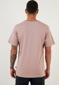 Tricou scurt cu mâneci scurte, de culoare roz deschis, cu decolteu rotund. Materialul pare moale, cu o textură netedă. Prezintă o etichetă subtilă a mărcii aproape de tiv.