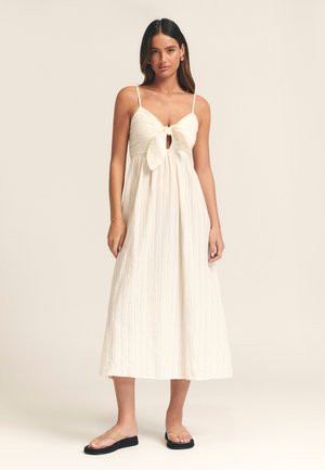 White, sleeveless maxi dress with a tie front and keyhole detail. Textured fabric with vertical stripes, worn with beige flip-flops.