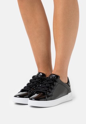 Nohy v lesklých černých nízkých teniskách s černými tkaničkami a bílými podešvemi stojící na jednobarevném světle pozadí.