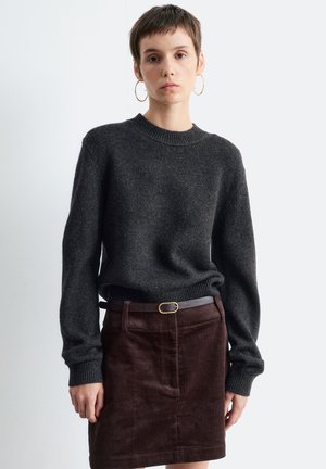 Woman with short hair wearing gold hoop earrings, dark grey sweater, brown corduroy skirt, and black belt, standing against a white background.
