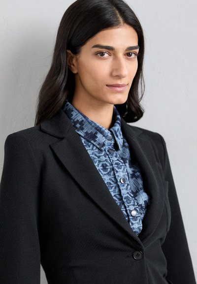 Svart blazer med en skräddarsydd design, lager på lager över en blå mönstrad skjorta med knäppning fram och krage.