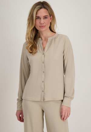 Vrouw met golvend blond haar, gekleed in een beige blouse met lange mouwen en knopen, en bijpassende broek, staand tegen een effen lichte achtergrond.