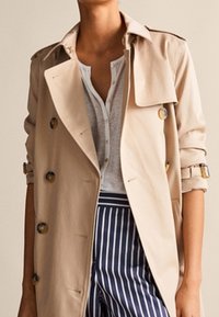 Trench-coat beige avec des détails de boutons sur un t-shirt henley blanc, associé à un pantalon rayé vertical bleu et blanc.