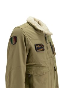 Giubbotto khaki con colletto in pelliccia sintetica, dotato di distintivi, inclusa una toppa blu "LEADER" e una toppa con bandiera multicolore. Tasche con zip.