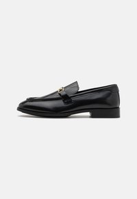 Mocassins en cuir noir avec un bout arrondi, des accents cousus, un petit talon et un détail en métal doré sur la sangle. Texture lisse dans l'ensemble.
