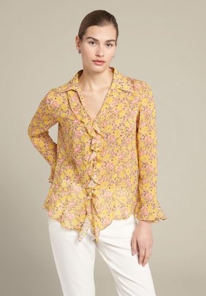 Geel bloem blouse met ruffles aan de voorkant, lange mouwen en een gekartelde zoom. Gemaakt van een lichte, doorschijnende stof met roze en groene accenten.