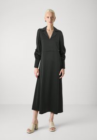 Black long-sleeve dress with a collared neckline, fitted bodice, and flowing skirt. Worn with light-colored block-heeled sandals.