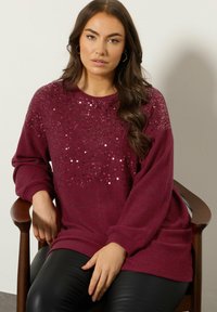 Bordowy sweter z ozdobami z cekinów w górnej części, luźny krój i długie rękawy; noszony na czarnych skórzanych spodniach.
