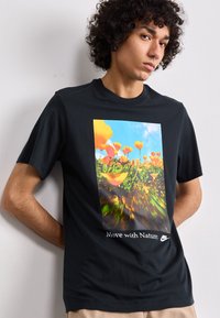 T-shirt preta com um gráfico floral colorido e o texto "Move with Nature" em branco, com o logótipo da Nike na parte inferior. Mangas curtas, decote redondo.