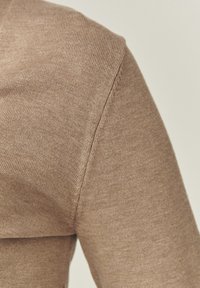 Pull en maille marron clair avec une coupe ajustée et une texture côtelée. La couture d'épaule est visible, mettant en valeur des détails de couture soignés.