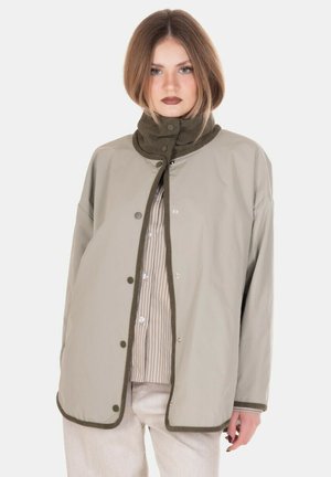 Giovane donna con capelli castano chiaro indossa giacca imbottita beige con bordi scuri sopra camicia a righe con bottoni e pantaloni chiari, sfondo neutro.