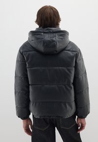 Chaqueta acolchada negra con capucha, que cuenta con secciones acolchadas y puños elásticos. El material parece suave y ligeramente brillante.