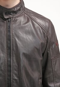 Brun läderjacka med hög krage, framzipper och sydda detaljer. Slät textur med en stilren, figursydd design.